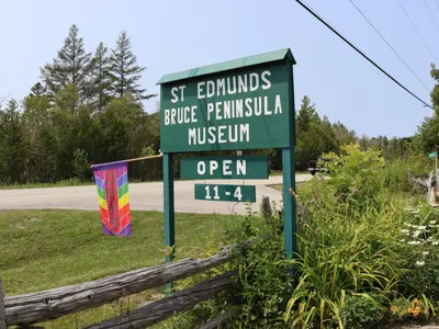 St. Edmunds Bruce Peninsula Museum roadside sign with “OPEN 11–4” and a rainbow flag, set against a rural, wooded backdrop.