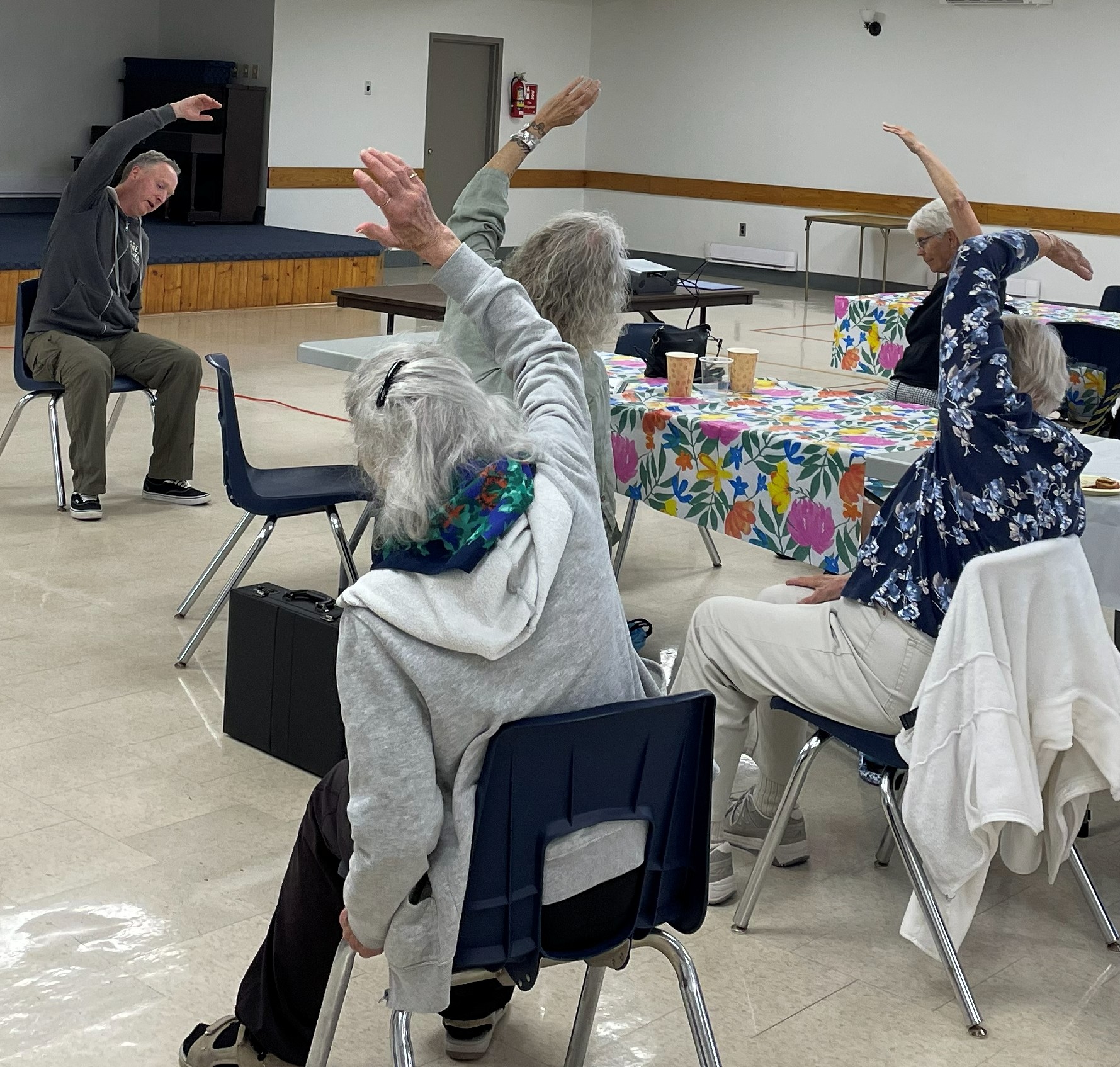 Shawn Radcliffe at front of room teaching 4 senior women chair yoga. All are seated.