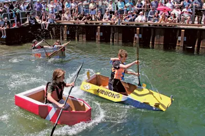 Children paddle homemade cardboard boats in a friendly race while a crowd watches from the dock.