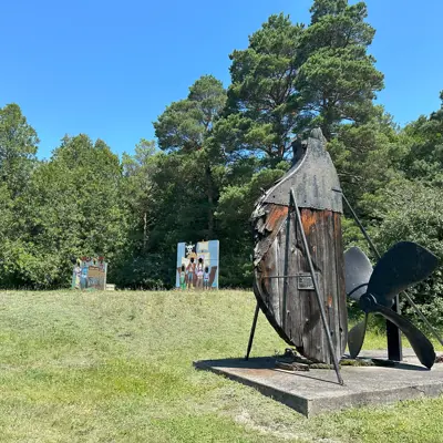 Wide view of a ship's rudder on display, outdoors in a grassy area with a rudder installation and surrounding trees.